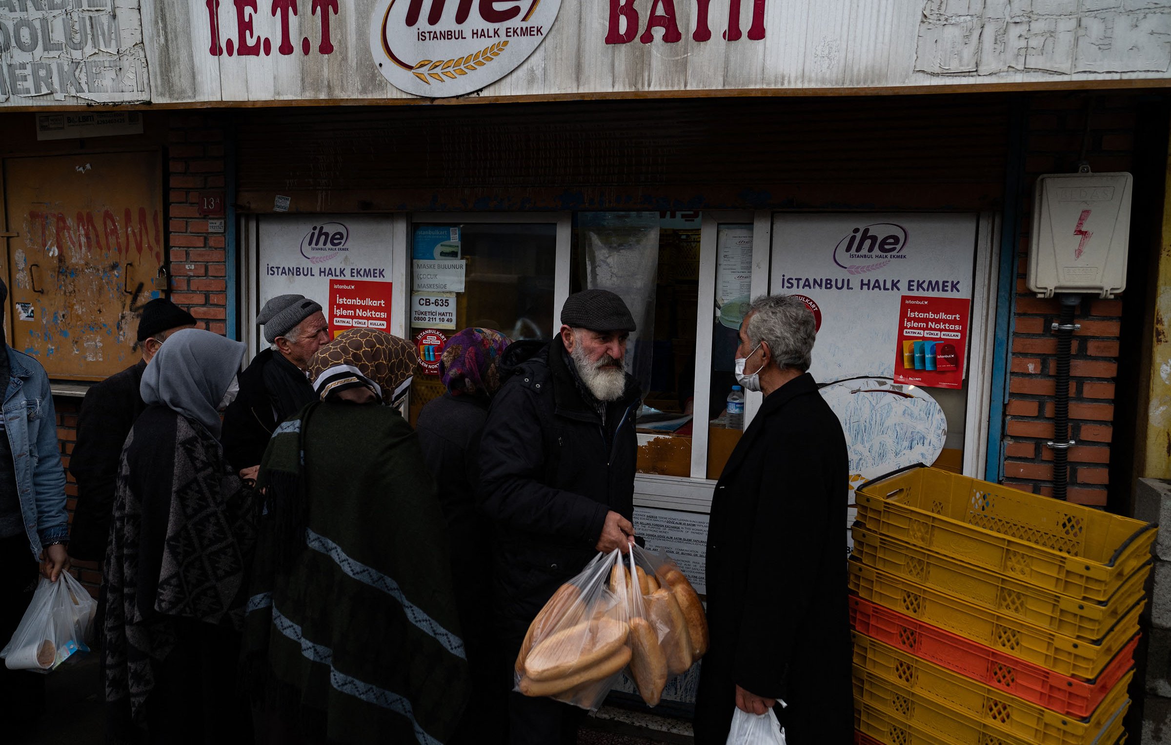 La imagen muestra a un grupo de personas en la calle, frente a una tienda. Algunos de ellos llevan bolsas en las manos, probablemente con pan. La escena sugiere una atmósfera de actividad comunitaria, ya que la gente está haciendo fila. Se pueden ver diferentes edades y estilos de ropa entre los presentes. Además, hay un letrero visible en la tienda que indica que se trata de un punto de venta de pan en Estambul.