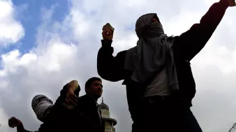 La imagen muestra a varias personas en un ambiente de tensión, lanzando objetos. Están parcialmente cubiertos con prendas que ocultan su rostro, lo que sugiere que están en una situación de protesta o conflicto. Al fondo, se puede ver una estructura que parece ser una mezquita, y hay alambre de púas en primer plano, indicando un entorno posiblemente conflictivo o restringido. El cielo está nublado, lo que añade una atmósfera sombría a la escena.