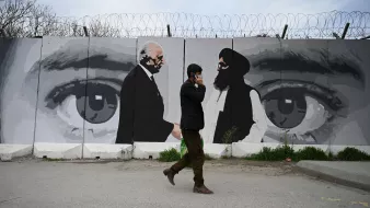 La imagen muestra a un hombre caminando por una calle, mientras habla por teléfono. Detrás de él hay un mural que representa dos figuras en blanco y negro, aparentemente en un diálogo. El mural incluye grandes ojos que observan la escena. Además, se pueden ver alambres de púa en la parte superior del muro que sostiene el mural, lo que sugiere un ambiente de tensión o seguridad. La iluminación es natural, indicando que es de día.