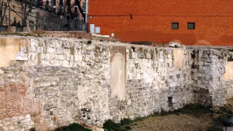 La imagen muestra una pared de piedra antigua, probablemente parte de una estructura histórica. La pared está compuesta de grandes bloques de piedra, algunos de los cuales están desgastados o dañados. En el centro, hay un espacio vacío en la pared que sugiere la existencia previa de una puerta o ventana. Al fondo, se puede ver un edificio de ladrillo rojo y un área de césped seco. Hay algunos restos de vegetación en la base de la pared, lo que añade un toque de naturaleza al entorno urbano.