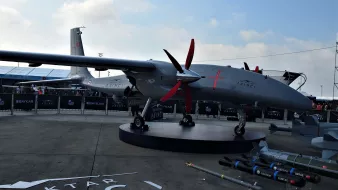 La imagen muestra un dron militar de la marca Baykar. Está expuesto en un evento, con un diseño aerodinámico y detalles en rojo. El dron tiene dos hélices rojas en el frente y un fuselaje gris, apoyado sobre una plataforma giratoria. Al fondo, se pueden observar otros drones y un ambiente que sugiere un evento de exhibición o industria militar. También hay misiles en primer plano, lo que indica su capacidad bélica.