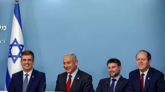 La imagen muestra a un grupo de cuatro personas sentadas alrededor de una mesa en un entorno oficial. Al fondo, se puede ver una bandera de Israel. Las personas parecen estar en un evento o una conferencia, y todos tienen expresiones serias pero amigables. Están vestidos de manera formal. La escena sugiere un contexto político o gubernamental.