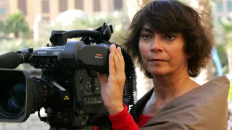 La imagen muestra a una mujer de cabello oscuro y rizado, que parece estar sosteniendo y operando una cámara de video profesional. Ella está mirando a la cámara con una expresión seria y concentrada. Viste una blusa color claro y tiene una capa o chal de un color más oscuro. Al fondo, se pueden ver edificios y un ambiente urbano. La escena sugiere que está en un evento o en el lugar de una grabación.