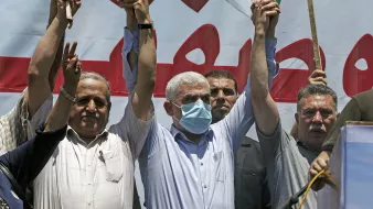En la imagen se puede ver a un grupo de hombres en una manifestación. Ellos levantan las manos, mostrando un gesto de unidad o celebración. Uno de los hombres en el centro lleva una máscara facial, lo que sugiere que la situación se desarrolla en un contexto donde se requieren medidas de salud. Detrás de ellos hay una pancarta o cartel grande, con texto en árabe y un fondo que parece estar relacionado con la causa por la que están protestando. La atmósfera parece ser de fervor y apoyo colectivo.
