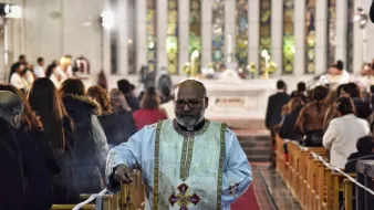 A priest in ornate vestments walks down an aisle in a church, surrounded by worshippers.