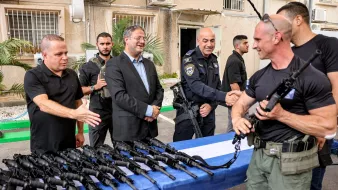 Men in uniform gather around rifles on a table, exchanging greetings and smiles.