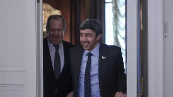 La imagen muestra a dos hombres entrando en una sala. Uno de ellos tiene una sonrisa amplia y está vestido con un traje oscuro y una corbata con un patrón. El otro hombre parece estar sonriendo también y lleva unas gafas. La decoración del fondo sugiere un ambiente formal y elegante, con luces suaves y detalles arquitectónicos. Ambos parecen estar en un momento de cordialidad y alegra.
