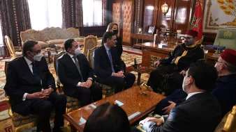 La imagen muestra una reunión en un ambiente formal y lujoso, posiblemente en un palacio. Hay varias personas sentadas alrededor de una mesa. Algunos de los asistentes llevan puesto un tapabocas, lo que sugiere que la reunión puede haber tenido lugar durante un periodo de preocupación por la salud pública, como la pandemia. En el centro de la escena, hay un individuo que parece ser una figura de autoridad, vestido con túnica y gorro tradicional. El interior está decorado con un estilo opulento, que incluye una alfombra elaborada y muebles de alta calidad. Las expresiones de los asistentes son serias, indicando la importancia del encuentro.