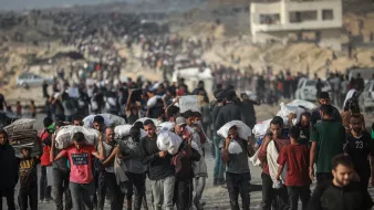 Un grupo de personas camina por un terreno árido, llevando sacos y luciendo cansados.
