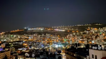 La imagen muestra una vista nocturna de la ciudad, iluminada por luces de diversos colores. Se pueden ver edificios y viviendas en la parte inferior, con antenas en los techos de algunas estructuras. En el fondo, hay colinas que también están iluminadas, y el cielo es oscuro, lo que resalta aún más las luces de la ciudad. Es un paisaje urbano que refleja la actividad y la vida nocturna del lugar.