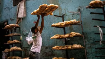Un hombre levanta canastas con pan en un ambiente de panadería. Las paredes son azules y desgastadas.
