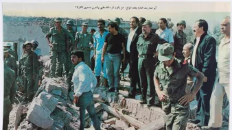 A group of military personnel and civilians gathers at a rubble site, surveying damage.