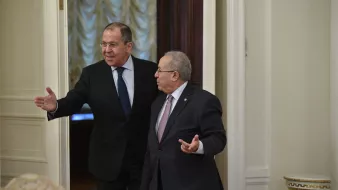 En la imagen se puede ver a dos hombres en un entorno formal, probablemente en una sala de reuniones o un despacho. Uno de ellos, a la izquierda, parece estar señalando o gesticulando con una mano, mientras que el otro, a la derecha, escucha y gira ligeramente su cuerpo hacia él. Ambos visten trajes formales y tienen expresiones serias. El ambiente parece elegante, con detalles en las paredes y una iluminación suave.