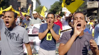 La imagen muestra a un grupo de personas participando en una manifestación. Los manifestantes parecen estar expresando su descontento, gritando y levantando las manos. En el fondo, se pueden ver pancartas y banderas, algunas de las cuales tienen un símbolo de mano levantada. El ambiente es de protesta, y la multitud está activa y unida en su mensaje. La escena se desarrolla en un entorno urbano, con edificios visibles en el fondo.