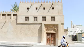 La imagen muestra un edificio de arquitectura tradicional, con paredes de color beige y detalles ornamentales en la fachada. La puerta principal es de madera tallada, y hay ventanas con rejillas decorativas. Al lado de la construcción, un ciclista vestido con ropa ligera y un casco pedalea por una calle pavimentada. El ambiente parece cálido, lo que sugiere un clima árido, típico de ciertas regiones. En el fondo, se pueden observar otros edificios y estructuras, que contribuyen a la sensación de un entorno urbano.