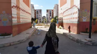 La imagen muestra una escena de una calle en un barrio residencial. En el primer plano, una mujer con un hiyab sostiene de la mano a un niño pequeño mientras caminan. Al fondo, se pueden ver edificios de apartamentos con pinturas decorativas en las paredes. El ambiente parece tranquilo y iluminado por la luz del día, y hay un pequeño espacio ajardinado en el centro, lo que sugiere un lugar comunitario.