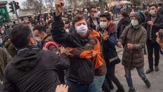 La imagen muestra a un grupo de personas en medio de una situación tensa. Se puede ver a un individuo en el centro, levantando el puño mientras es sujetado por otro, aparentemente en una escena de protesta o manifestación. La mayoría de las personas en la imagen llevan mascarillas. El ambiente parece ser urbano, con un semáforo y algunos edificios de fondo. La expresión de los individuos sugiere emociones intensas, probablemente relacionadas con el conflicto o la agitación social.