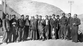 La imagen muestra un grupo de hombres de pie en un entorno rural, posiblemente en una zona montañosa. Están vestidos con ropas tradicionales y de trabajo, algunos con boinas. Hay también un perro que se encuentra entre ellos. La atmósfera es de camaradería, y el paisaje detrás sugiere una época pasada, posiblemente de principios del siglo XX. Los rostros reflejan diferentes expresiones, mientras que la composición transmite una sensación de unión entre los miembros del grupo.