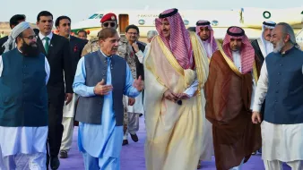 En la imagen, se puede ver a un grupo de hombres en un evento formal. Uno de ellos, vestido con una túnica azul, parece estar conversando animadamente mientras camina. Los demás, vestidos con trajes tradicionales y de gala, incluyen a personas con vestimenta árabe y otros trajes formales. Están rodeados de un ambiente elegante, con un jet privado y una alfombra morada en el fondo. La escena sugiere una ocasión diplomática o una visita de estado.