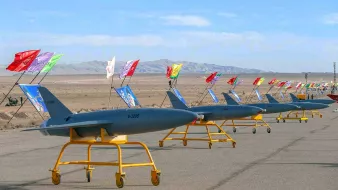 La imagen muestra una fila de drones en una pista desértica. Estos drones están montados sobre soportes amarillos y llevan banderas de diferentes colores. Al fondo, se puede ver un paisaje montañoso y un cielo despejado. La escena parece estar relacionada con una exhibición o un evento militar.
