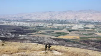 La imagen muestra un paisaje vasto y montañoso, con colinas y valles amplios. En primer plano, se pueden ver dos personas de pie, posiblemente soldados, observando el paisaje desde una cima. El área está cubierta de hierba seca y destaca un amplio valle con campos de cultivo en diferentes tonos de verde y amarillo. En el fondo, se aprecian montañas que se extienden hasta el horizonte bajo un cielo despejado.