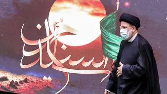 La imagen muestra a un hombre vestido con un atuendo tradicional islámico, que incluye un turbante negro y una túnica oscura. Está usando una mascarilla facial. Detrás de él, se puede ver un fondo colorido con un diseño que incluye una luna y una bandera, junto con un texto en árabe. La atmósfera parece estar relacionada con un evento significativo cultural o religioso.