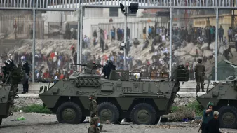 La imagen muestra un contexto de tensión, probablemente en una zona de conflicto o crisis. Se pueden ver vehículos militares blindados en la parte frontal, con soldados a bordo y personal militar en el entorno. Al fondo, hay una multitud de personas que parecen estar frente a una valla alta, indicando una separación entre diferentes grupos. El ambiente es de intensa actividad, posiblemente con indicios de enfrentamientos o una situación de disturbio. La escena evoca un fuerte sentido de vigilancia y seguridad militar.