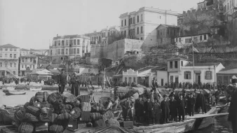 La imagen muestra un puerto animado en un contexto histórico, con un barco de madera cargado de bultos y barriles en primer plano. Un grupo de personas, posiblemente trabajadores o comerciantes, está de pie sobre la embarcación. Al fondo, se puede ver una serie de edificios y una muralla de piedra que se eleva, sugiriendo la presencia de una ciudad costera. La escena tiene un aspecto antiguo, lo que sugiere que fue tomada en una época pasada, posiblemente a finales del siglo XIX o principios del XX. Las actividades portuarias y el diseño arquitectónico transmiten una sensación de vida cotidiana en el puerto.