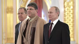 La imagen muestra a tres hombres de pie en un entorno ceremonial. El hombre a la izquierda lleva un traje oscuro y tiene una expresión seria. En el centro, el segundo hombre viste una prenda con un diseño decorativo y también parece estar en una postura formal. A la derecha, se encuentra otro hombre, vestido con un traje oscuro y corbata, que observa al frente. El fondo tiene un ambiente lujoso con detalles dorados y alusiones a un contexto oficial.