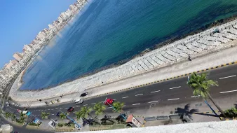 La imagen muestra una vista aérea de una costa bien iluminada. Se puede ver una carretera que corre paralela a la playa, con varios vehículos transitando. A lo largo de la playa hay palmeras, y el agua del mar es de un color azul claro. Al fondo, se pueden observar edificios que forman parte de un área urbana, y el cielo está despejado, lo que sugiere un día soleado.