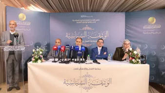 La imagen muestra a un grupo de cuatro personas sentadas en una mesa de prensa durante un evento. Hay micrófonos de diferentes medios de comunicación frente a ellos, y el ambiente parece ser formal. La persona de pie, a la izquierda, parece estar dando un discurso o presentación. El fondo incluye un cartel con texto en árabe, y hay arreglos de flores sobre la mesa. La escena sugiere que se está llevando a cabo una conferencia o reunión importante.