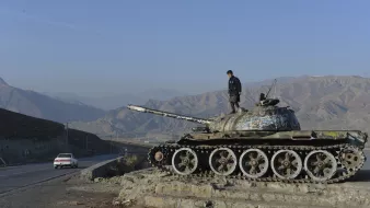 La imagen muestra a un joven de pie sobre un tanque de guerra, que parece estar fuera de uso. El tanque tiene un acabado desgastado y está cubierto de óxido y pintura descascarada. Al fondo, se pueden ver montañas cubiertas de vegetación y un cielo despejado. A un lado, hay una carretera donde se observa un automóvil blanco en movimiento. La escena transmite una sensación de desolación y el contraste entre la guerra y la paz.