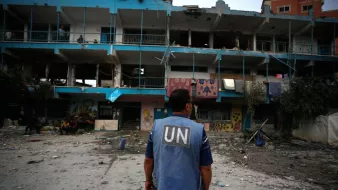 The image depicts a person wearing a blue vest labeled "UN," standing in front of a damaged building. The structure shows signs of destruction, with broken windows and debris scattered around the ground. There are individuals visible in the background, and the overall scene conveys a somber atmosphere, likely in a conflict or disaster-stricken area.