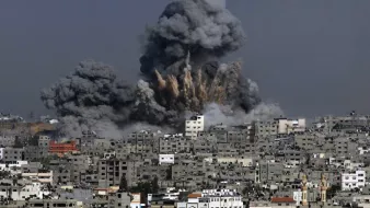 La imagen muestra una explosión en un área urbana, donde se elevan grandes nubes de humo y escombros hacia el cielo. Las estructuras de edificios en la parte inferior parecen estar dañadas, y el entorno refleja un ambiente de conflicto o desastre. La atmósfera es de caos, con el cielo grisáceo debido al humo de la explosión.