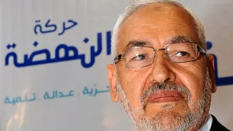 La imagen muestra a un hombre mayor con barba y gafas, mirando hacia un lado. Lleva una camisa clara y un saco oscuro. Al fondo se puede ver un logo que incluye una estrella roja y texto en árabe y en español que parece estar relacionado con un movimiento político o una organización. El ambiente sugiere un contexto formal o de conferencia.