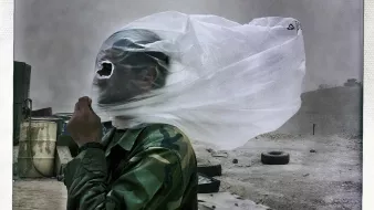 La imagen muestra a un hombre vestido con un uniforme de camuflaje, protegiéndose de una tormenta de arena. Su rostro está cubierto con una tela transparente que parece ser una especie de máscara o protección. El entorno se ve polvoriento y desolado, con algunos objetos y neumáticos dispersos en el área. La escena transmite una sensación de dificultad y adversidad debido a las condiciones climáticas.