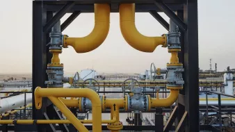 La imagen muestra una estructura industrial, probablemente de una planta de procesamiento de petróleo o gas. Hay varias tuberías de color amarillo que se conectan entre sí, junto con válvulas y otros elementos metálicos. El fondo sugiere un paisaje industrial con tanques y equipos, lo que indica un entorno de trabajo relacionado con la energía o la petroquímica. La escena parece ser al aire libre, bajo un cielo claro.