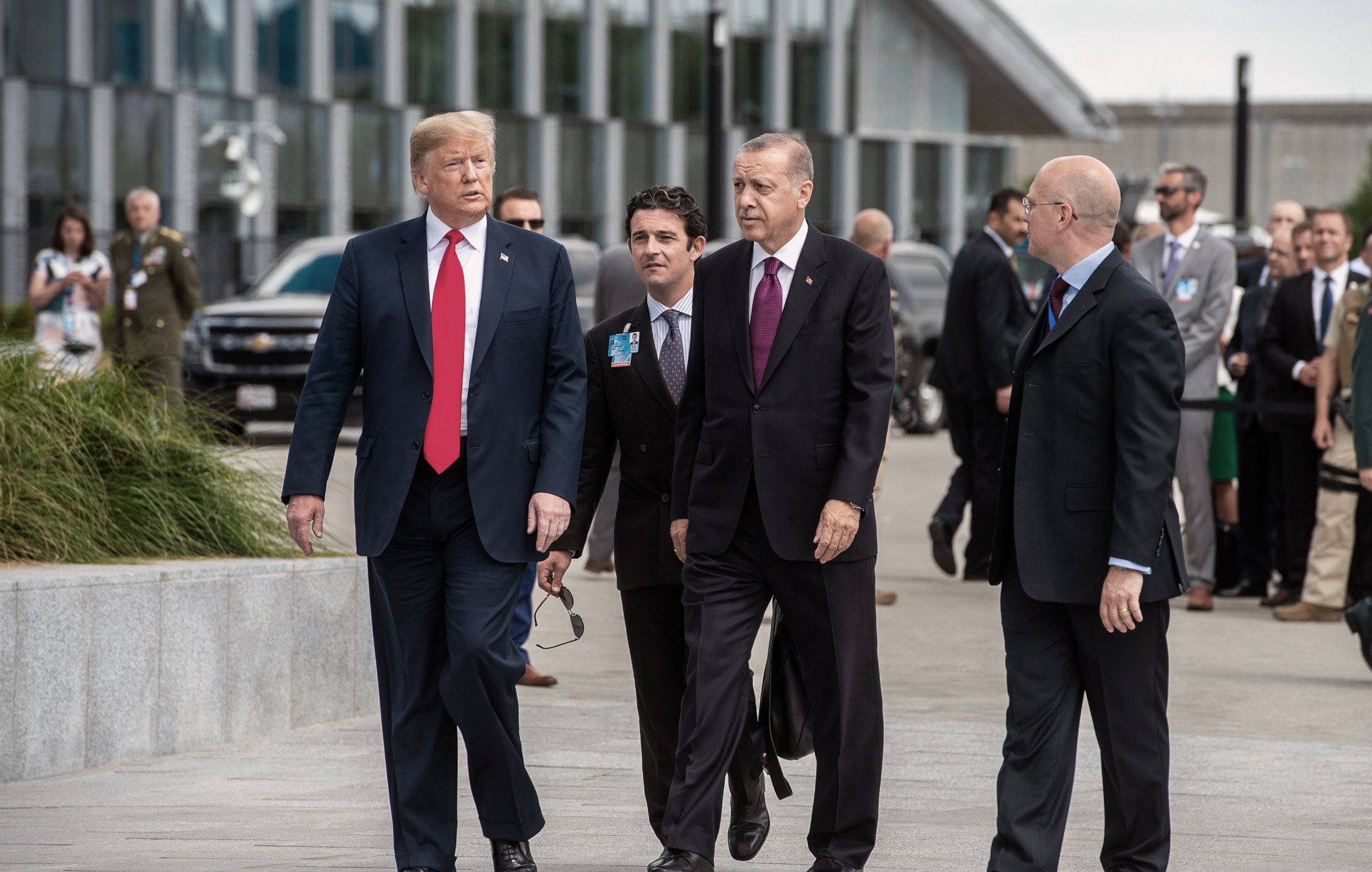 در این تصویر، چند مرد در حال پیادهروی هستند. یکی از آنها که لباس رسمی پوشیده و کلاهی تیره به سر دارد، به سمت دیگران نگاه میکند. شخص دیگری که در سمت چپ او قرار دارد، کت و شلوار و یک کراوات قرمز به تن دارد. دو مرد دیگر نیز به آنها ملحق شدهاند و ظاهراً در حال گفتگو هستند. پسزمینه، شامل ساختمانهای مدرن و تعدادی از افراد دیگر در حال حرکت است. جو کلی تصویر نشاندهنده یک ملاقات رسمی است.