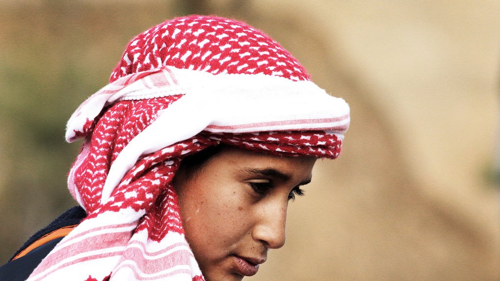 در این تصویر، یک فرد با چهرهای متعلق به سن جوانی دیده میشود. او یک کلاهی با طرح قرمز و سفید بر سر دارد که به شیوهای سنتی پوشیده شده است. چهرهاش به سمت پایین متمایل است و نگاهی تفکرآمیز دارد. پسزمینه تصویر مبهم است و توجه بیشتر بر روی شخص متمرکز شده است. این تصویر حس آرامش و تمرکز را منتقل میکند.