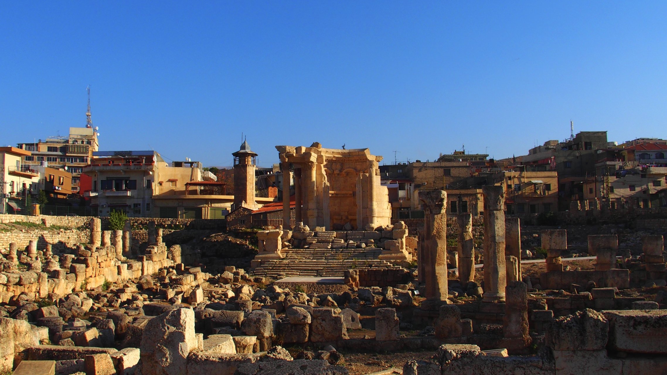 این تصویر نمایی از یک مکان باستانی را نشان میدهد که شامل خرابههای ستونها و دیوارهای قدیمی است. در پسزمینه، چند ساختمان مدرنتر دیده میشود که نشاندهنده ترکیبی از تاریخ با معماری معاصر است. آسمان آبی و صاف است و نور خورشید بر روی سنگها میتابد که حس زیبایی و آرامش را به تصویر میبخشد. اجزای باستانی این مکان نشاندهنده تاریخ غنی و فرهنگی آنجا هستند.