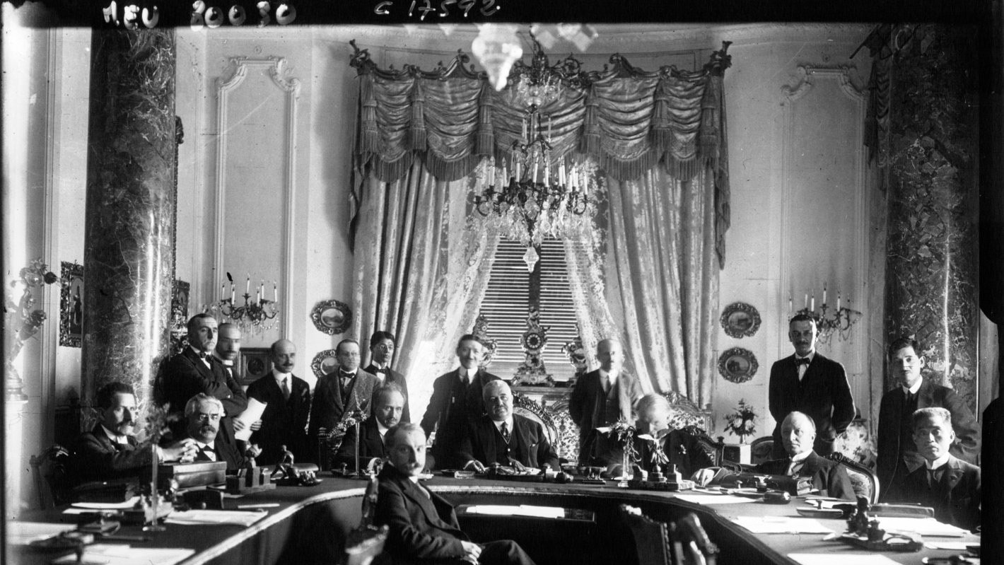 این تصویر، یک جلسه یا کنفرانس تاریخی را نشان میدهد که در آن گروهی از مردان در یک اتاق بزرگ و با طراحی تجملاتی گرد هم آمدهاند. در مرکز تصویر، یک مردی با لباس رسمی نشسته است و دیگر افراد دور او ایستاده و نشستهاند. دکوراسیون اتاق شامل پردههای بلند، لوستر و تزئینات دیواری است که به فضای جلسه شکوه و جلال خاصی میبخشد. این تصویر نشاندهنده یک رویداد مهم و رسمی در تاریخ است.