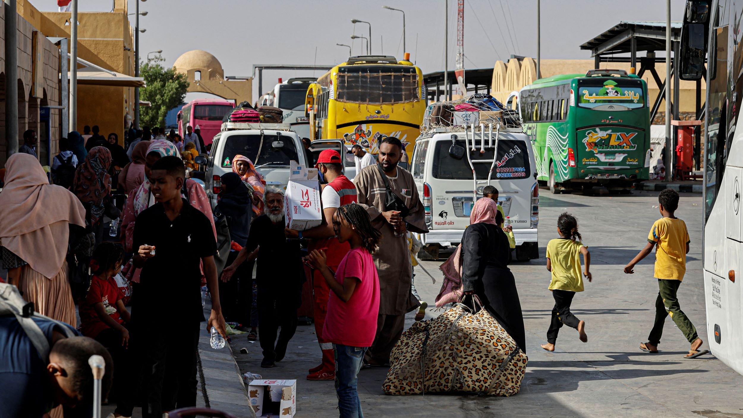 در تصویر، یک ایستگاه اتوبوس یا ترمینال مشاهده میشود که در آن تعدادی اتوبوس در حال توقف هستند. مردم زیادی در حال رفت و آمد هستند، برخی در حال حمل چمدان و بارهای خود هستند. در میان این افراد، کودکان نیز دیده میشوند که در حال بازی یا حرکت هستند. برخی از افراد در حال استراحت و نشستن بر روی ساکهای خود هستند. فضای ترمینال به نظر شلوغ و پرجنب و جوش میآید و آفتاب روشن در حال تابش است.