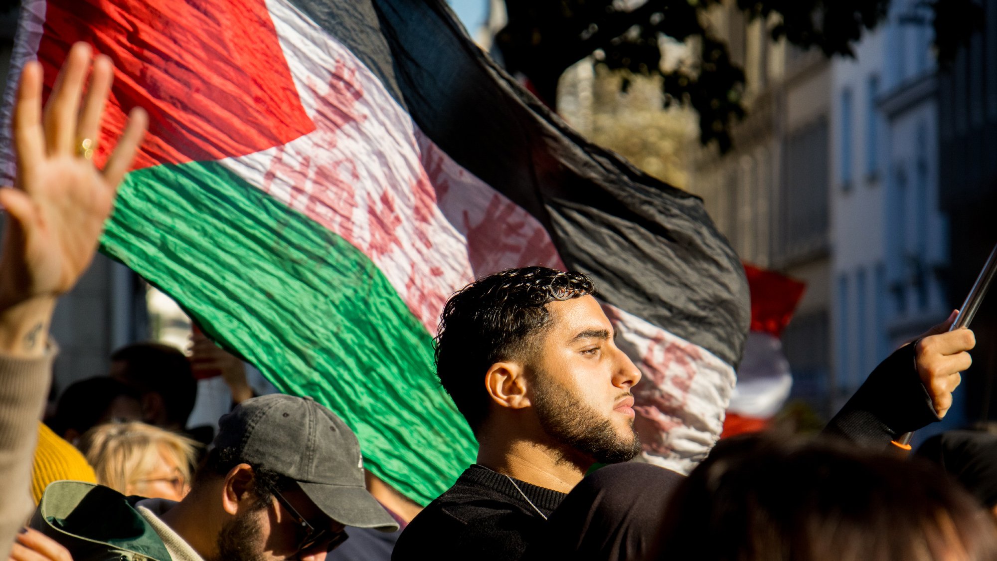 در این تصویر، گروهی از افراد در حال برگزاری یک تظاهر یا تجمع هستند. یک مرد جوان در جلو تصویر دیده میشود که به پرچم کشور خود (که احتمالاً پرچم فلسطین است) افتخار میکند. او با جدیت به جلو نگاه میکند. در پسزمینه، تعدادی از افراد دیگر نیز حضور دارند که برخی از آنها کلاه به سر دارند و به نظر میرسد که در حال حمایت از یک هدف مشترک هستند. نور آفتاب به تصویر زیبایی لطافتی میبخشد و فضای تجمع را گرم و زنده نشان میدهد.