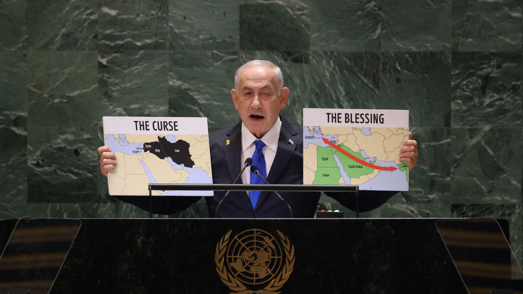 در این تصویر، یک مرد در حال صحبت در پشت یک تریبون است. او دو نقشه را در دست دارد که بر روی یکی از آنها نوشته شده "نفرین" و بر روی دیگری "برکت". مرد به سمت حضار اشاره میکند و به نظر میرسد در حال بیان یک موضع سیاسی یا اجتماعی مهم است. پسزمینه تریبون نشاندهنده نماد سازمان ملل متحد است.