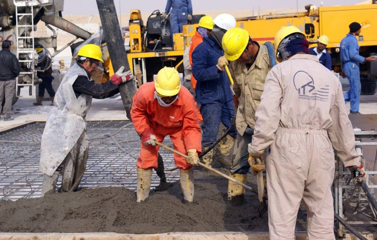 در این تصویر گروهی از کارگران مشغول به کار در یک پروژه ساختمانی هستند. آنها کلاه ایمنی به سر دارند و لباسهای کار پوشیدهاند. چند نفر در حال ریختن بتن و استفاده از تجهیزات مختلف هستند. محیط کار شلوغ و صنعتی به نظر میرسد و ماشینآلاتی در پسزمینه دیده میشود. کارگران با جدیت و همکاری در حال انجام وظایف خود هستند.