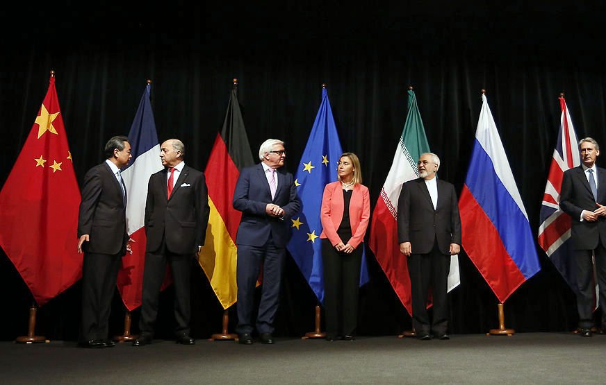 در این تصویر، گروهی از نمایندگان کشورهای مختلف در یک جلسه رسمی ایستادهاند. پرچمهای چین، آلمان، اتحادیه اروپا، ایران، روسیه، بریتانیا و ایالات متحده در پسزمینه حضور دارند. هر یک از افراد در لباسهای رسمی و با حالتهای متفاوتی در صف ایستادهاند، که نشاندهنده اهمیت و جدیت جلسه است. به نظر میرسد آنها در حال گفتوگو و تعامل با یکدیگر هستند.