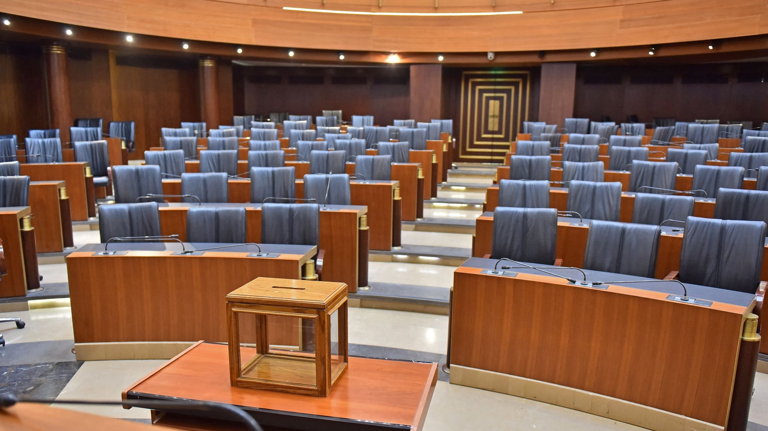 این تصویر نمایی از یک سالن جلسات یا تالار کنفرانس را نشان میدهد. صندلیهای چوبی و چرمی مشکی با فاصله منظم در ردیفهای متعدد چیده شدهاند. در وسط تالار یک میز چوبی با یک جعبه شیشهای کوچک وجود دارد. نورپردازی سالن به نظر نسبتاً ملایم است و دیوارها و سطوح چوبی به فضا حس رسمی و معتبر میدهند.