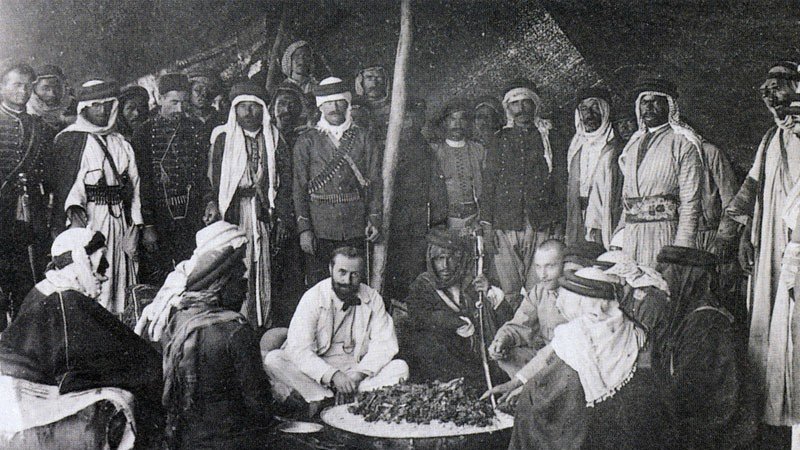 این تصویر تاریخی گروهی از مردان را در یک محیط سنتی نشان میدهد. آنها در یک چادر یا فضای باز گرد هم آمدهاند و به نظر میرسد در حال تهیه یا صرف غذا هستند. چند نفر از آنها لباسهای سنتی عربی به تن دارند، از جمله عبا و headscarf (کشیه). در مرکز تصویر، چند نفر در حال نشستن و گفتگو هستند و سایرین ایستاده یا نشسته در اطراف آنها قرار دارند. فضای تصویر گرم و صمیمی به نظر میرسد و نمایانگر فرهنگی غنی و سنتهای اجتماعی است.