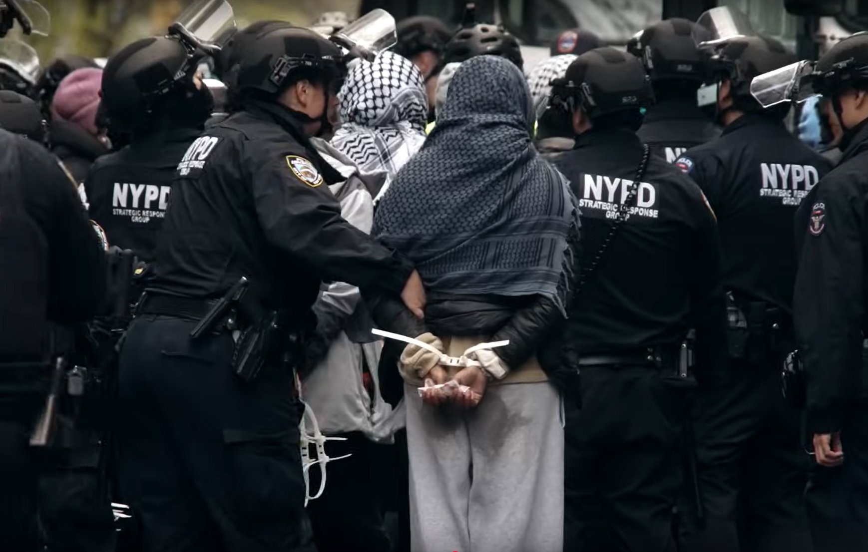 تصویر یک تجمع اعتراضی است که در آن پلیسها با لباس سیاه و کلاهخود، به یک فرد که دستبند به دستانش زده شده است، نزدیک میشوند. این فرد لباسهای غیررسمی و یک پوشش روی سر دارد. محیط به نظر شلوغ و پر از پلیسهای دیگر است.