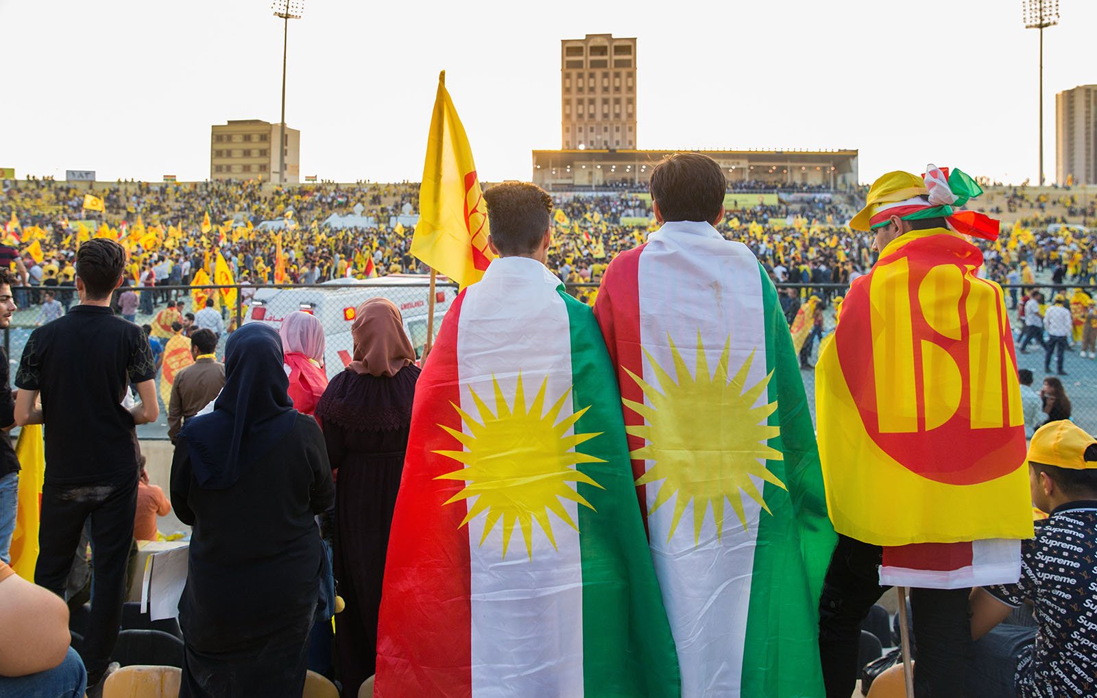 در این تصویر، گروهی از مردم در یک استادیوم جمع شدهاند. در پیشزمینه، سه نفر دیده میشوند که در حال تماشا هستند و روی شانههایشان پرچمهای کردستان و سمبلهای مرتبط را دارند. سایر تماشاگران در دور و اطراف نیز حضور دارند و فضا به شدت رنگارنگ و شاداب به نظر میرسد. اکثر افراد در جمعیت لباسهای زرد و قرمز پوشیدهاند و جو به وضوح نمایشگر شور و هیجان است. غروب خورشید، نور ملایمی بر روی جمعیت میتاباند و فضایی دوستانه ایجاد کرده است.